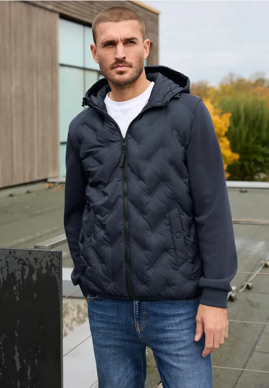 Un homme en doudoune sombre, t-shirt blanc et jean bleu se tient sur une terrasse avec bâtiment en arrière-plan.