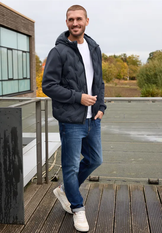 Homme en doudoune sombre, jean et baskets blanches, debout sur une terrasse urbaine devant un paysage arboré.