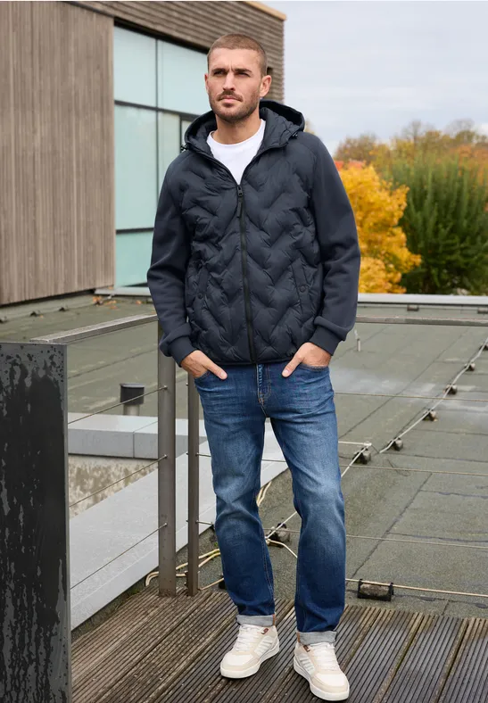 Homme sur balcon, manteau matelassé bleu, jeans et baskets blanches, paysage d'automne avec arbres colorés.