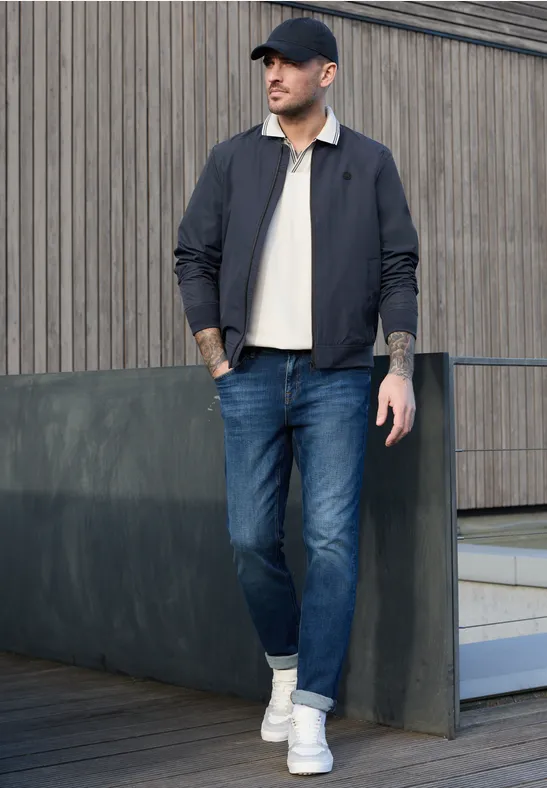 Een man in een zwarte jas, spijkerbroek en witte sneakers staat voor een modern gebouw.