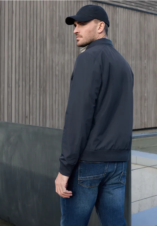 Een man in een zwart jack, spijkerbroek en pet staat voor een houten gevel.