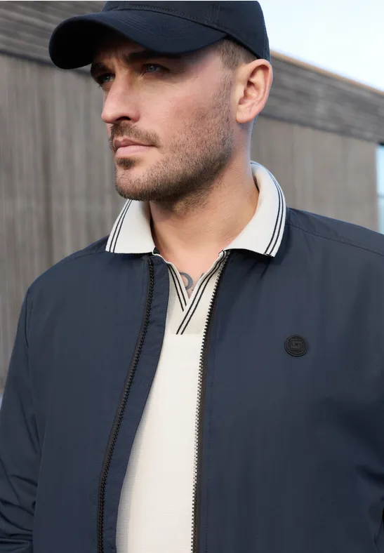 Een man draagt een donkerblauwe jas op een licht poloshirt voor een houten wand.