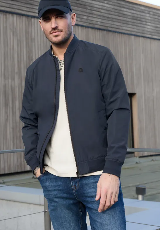Een man in donkerblauw jasje, wit T-shirt en spijkerbroek staat voor een moderne houten gevel.