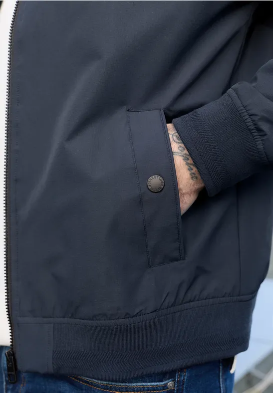 Close-up van een donkerblauwe jas met rits, zak en deels zichtbare getatoeëerde hand.