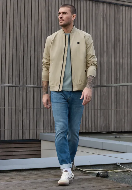 Man in beige jas, turquoise T-shirt, blauwe jeans en witte sneakers voor een houten wand.