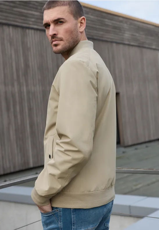 Een man in een beige jas staat zijwaarts met handen in zakken voor een houten wand.