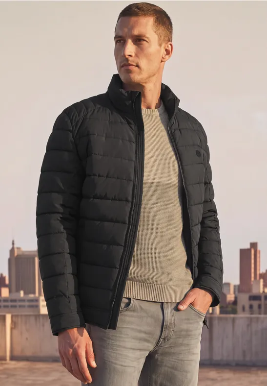 Mann in schwarzer Steppjacke und Jeans auf Dachterrasse vor Stadtsilhouette.