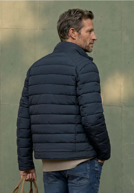 Ein Mann in dunkelblauer Steppjacke und Jeans steht seitlich vor einer grünen Wand.