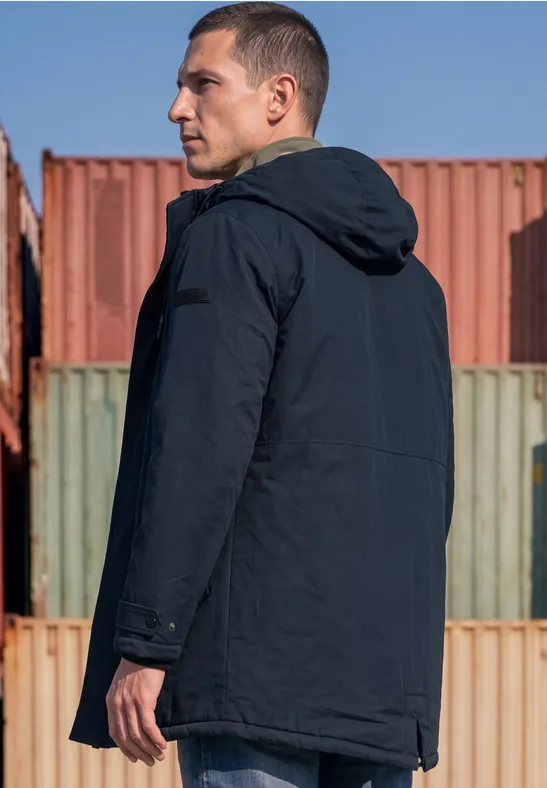 Ein Mann in einem dunklen Parka mit Kapuze steht seitlich vor bunten Containern.
