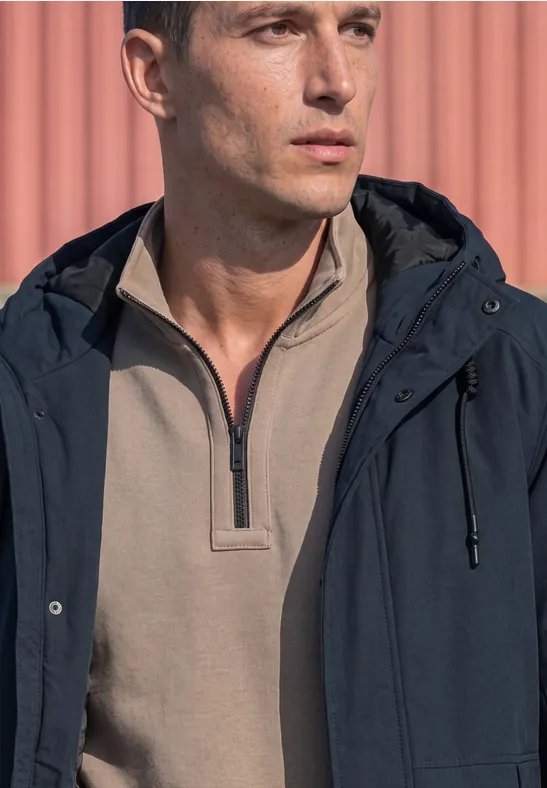 Ein Mann mit beigem Halbzip-Shirt und dunkelblauer Jacke steht vor einer rosa Wand.
