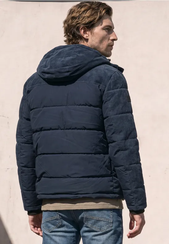 Een man draagt een blauwe gewatteerde jas met capuchon, gezien van achteren voor een lichte muur.