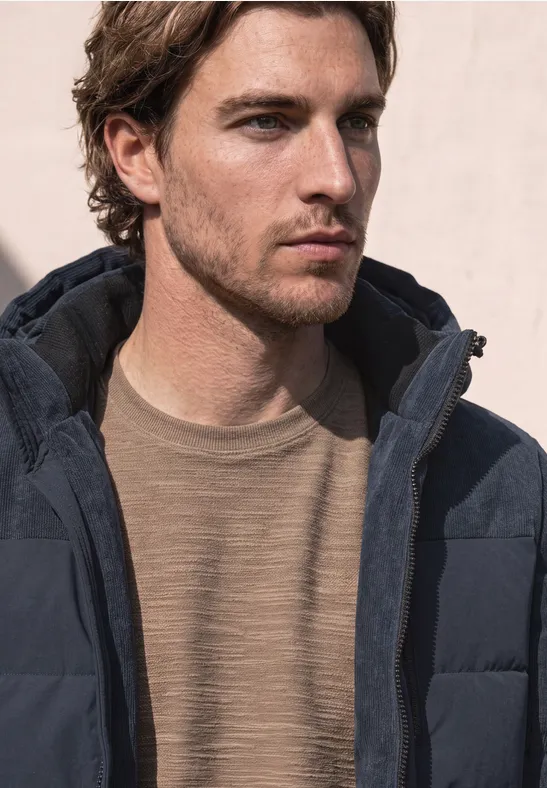 Een man met een donzen jas en een beige trui kijkt naar rechts voor een lichte achtergrond.