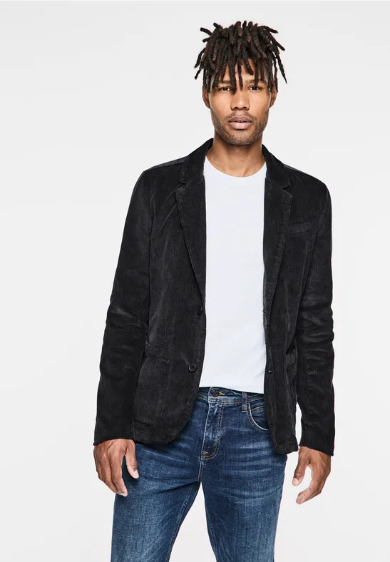 Man met zwarte dreadlocks, zwart colbert, wit shirt en spijkerbroek.