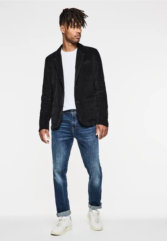 Man met zwart colbert, wit shirt, spijkerbroek en sneakers op witte achtergrond.