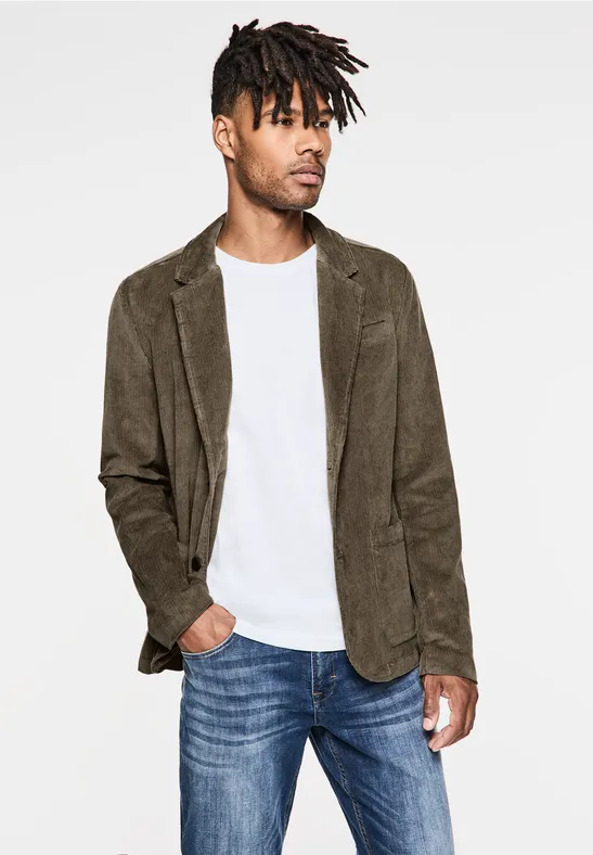 Man met bruin corduroy colbert, wit shirt en spijkerbroek tegen witte achtergrond.