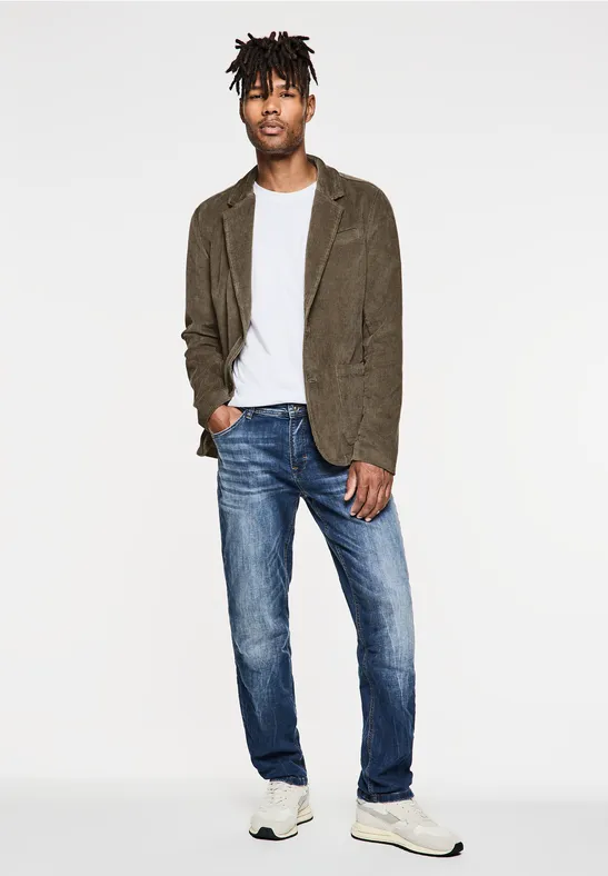 Man met wit T-shirt, jeans, sneakers en bruin colbert poseert relaxed.