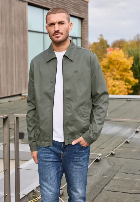 Ein Mann steht lässig auf einem Dach mit grüner Jacke, weißem T-Shirt und Jeans vor herbstlicher Kulisse.