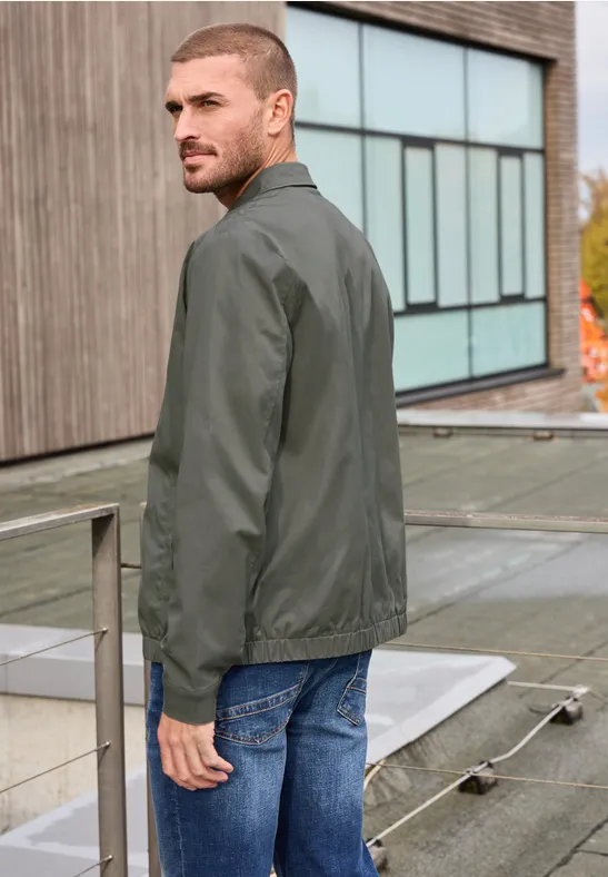 Ein Mann in grüner Bomberjacke und Jeans steht vor einem modernen Gebäude.