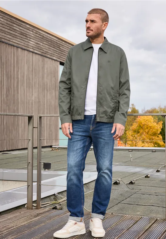 Ein Mann steht auf einem Dach mit grüner Jacke, weißem Shirt, blauen Jeans und weißen Sneakern.