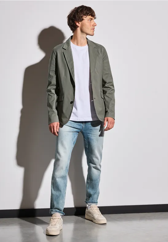 Man in grijs colbert, wit shirt en spijkerbroek voor donkere achtergrond.