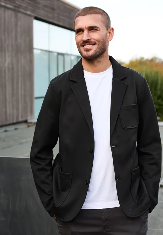 Een man in een zwart jasje en wit T-shirt staat voor een modern houten gebouw.
