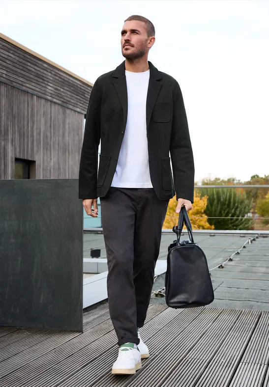 Man in zwart jasje, wit T-shirt, zwarte broek en witte sneakers, draagt een zwarte tas op een houten terras.