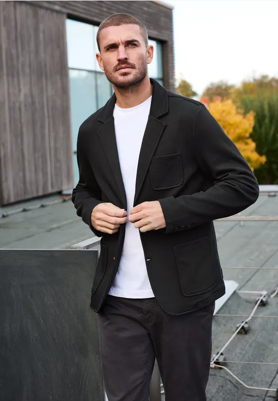 Een man op een dak, in een zwarte blazer, wit t-shirt en donkere broek met herfstige achtergrond.