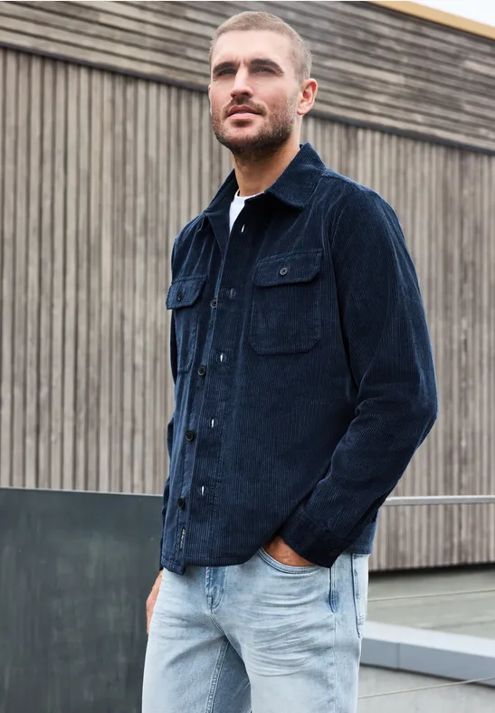 Man met kort haar in donkerblauwe corduroy jas en lichte jeans voor een houten wand.