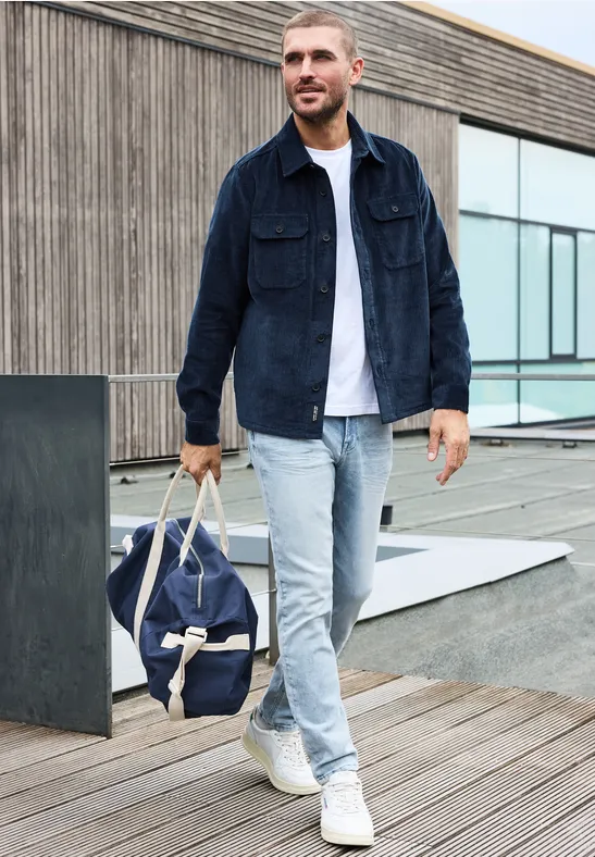 Een man in spijkerjack, witte T-shirt en lichte spijkerbroek loopt over een houten terras met een reistas.