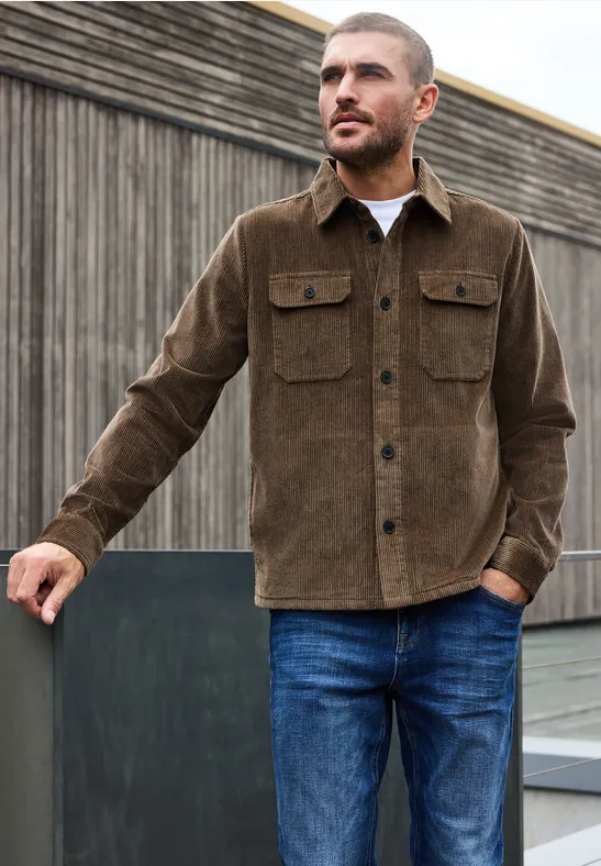 Een man leunt tegen een houten wand, draagt een bruin corduroy jack en een blauwe jeans.