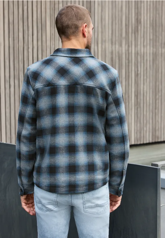 Ein Mann trägt von hinten gesehen ein blau-schwarzes Karohemd und helle Jeans vor einer Holzfassade.