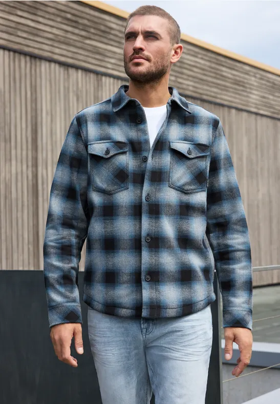 Ein Mann trägt ein kariertes blaues Hemd und hellblaue Jeans vor einer Holzwand.
