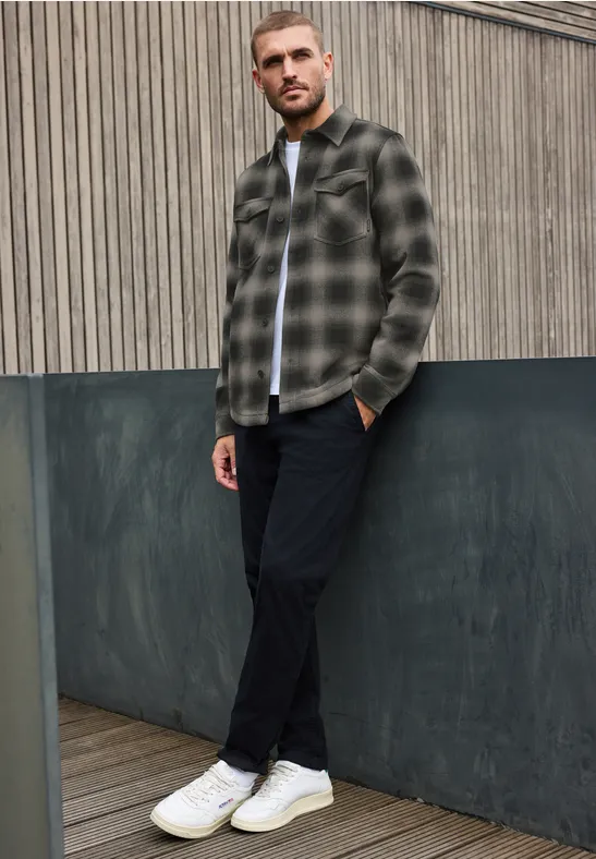 Ein Mann lehnt vor grauer Fassade, trägt kariertes Hemd, schwarze Hose und weiße Sneaker