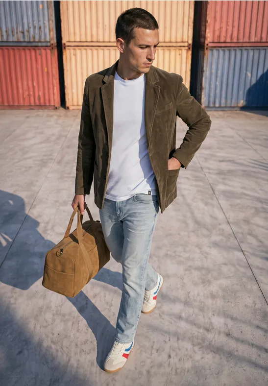 Eleganter Mann mit Sakko, weißem T-Shirt und Jeans trägt eine Reisetasche vor bunten Containern.