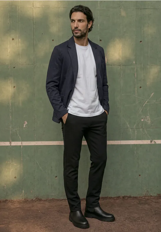 Een man met blazer, wit T-shirt, zwarte broek en laarzen staat voor een groene muur.