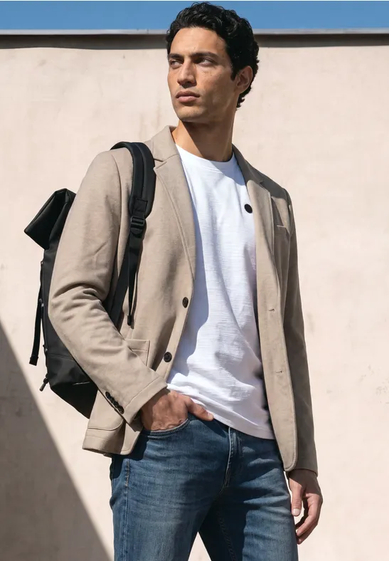 Een man staat voor een muur, draagt een beige jasje, een wit T-shirt, blauwe jeans en een rugzak.