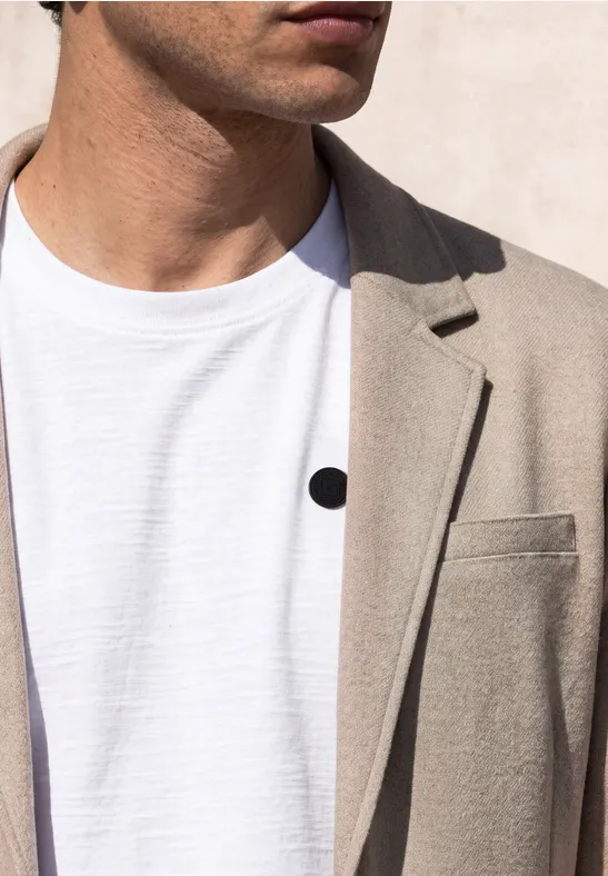 Een man draagt een beige colbert over een wit t-shirt met een klein zwart puntje op de borst.