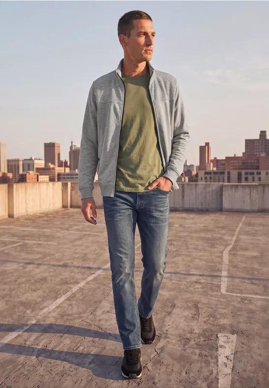 Ein Mann in Jeans, grünem Shirt und grauer Jacke läuft über ein Parkdeck mit Stadtkulisse im Hintergrund.