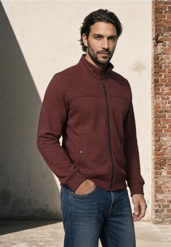 Un homme aux cheveux foncés et barbe porte un jean et une veste polaire bordeaux devant un mur.