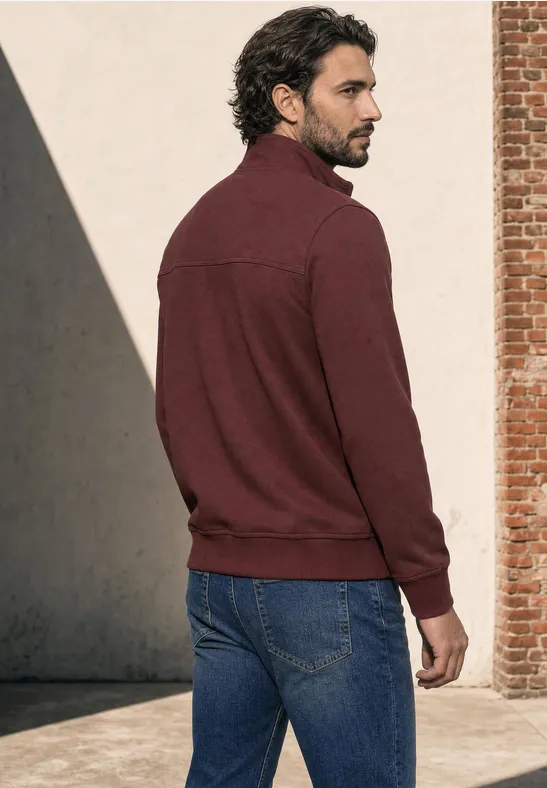 Un homme en pull bordeaux et jean se tient devant un mur en brique, regard de profil.