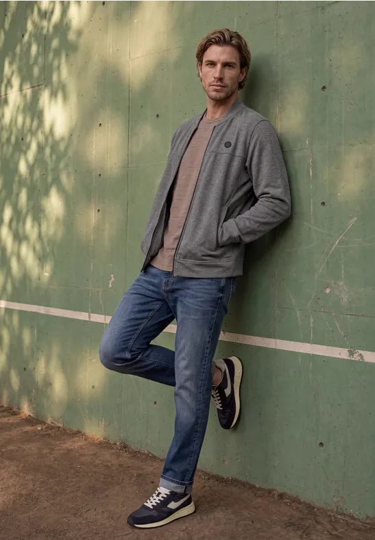 Een jonge man leunt ontspannen tegen een groene muur, gekleed in jeans, sneakers en een grijs jasje buiten.
