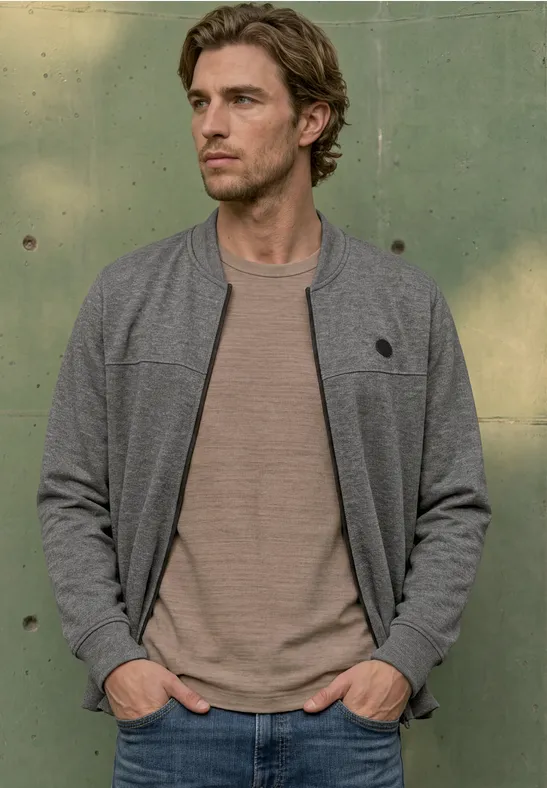Een man in een grijs bomberjack en beige t-shirt, handen in zakken, tegen een groene muur leunend.