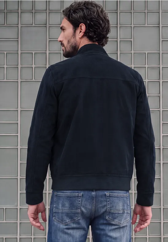 Ein Mann steht mit dem Rücken zur Kamera, trägt eine schwarze Jacke und Jeans vor grauer Wand.