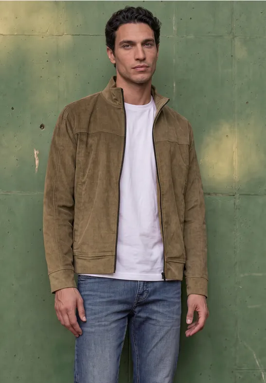Un jeune homme porte une veste en velours côtelé marron, un t-shirt blanc et un jean, devant un mur vert.