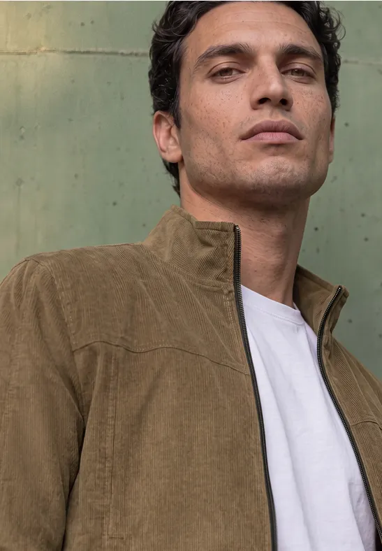 Jeune homme portant une veste en velours côtelé marron, col montant, t-shirt blanc, devant un mur vert pâle.