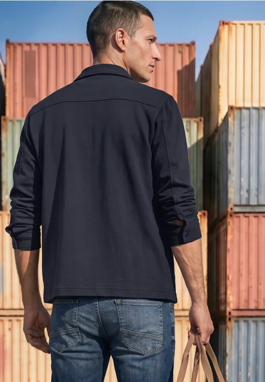 Ein Mann steht von hinten in dunkler Jacke und Jeans vor bunten Containern und hält eine Tasche.
