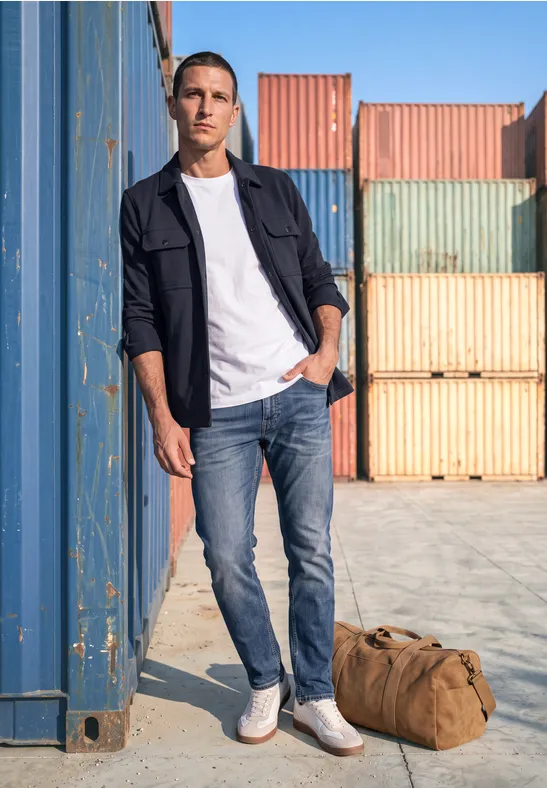 Ein Mann steht in einem Containerhafen, lehnt an einem bunten Container, trägt Jeans, weißes T-Shirt, Jacke und Tasche.