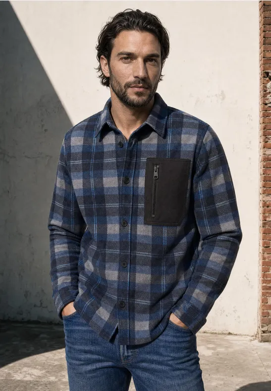 Man in blauw geruit flanellen overhemd en jeans, handen in zakken, voor een muurtje.