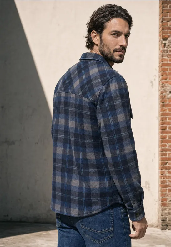 Een bebaarde man in een blauwe geruite blouse staat zijwaarts voor een muur en kijkt weg.