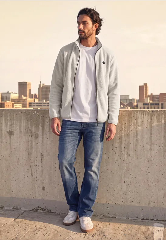 Ein Mann in weißem Hoodie, T-Shirt, Jeans und Sneakern steht auf einem Betonparkdeck mit Stadtblick.